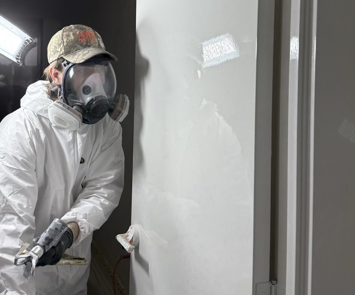 Worker in protective gear applying wall primer with a paint sprayer indoors.
